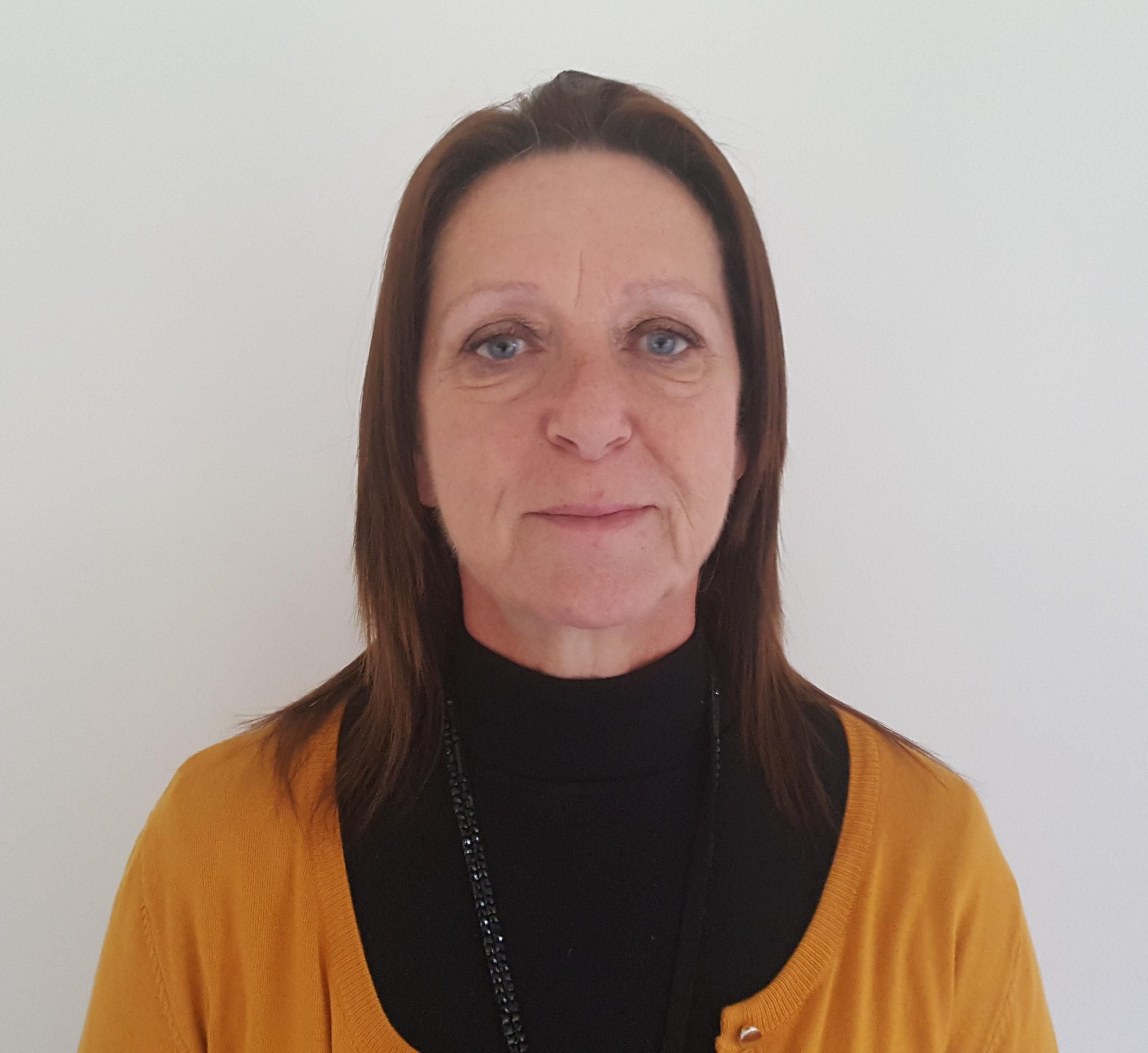 Woman looking at camera, with brown hair, blue eyes, black top and yellow cardigan