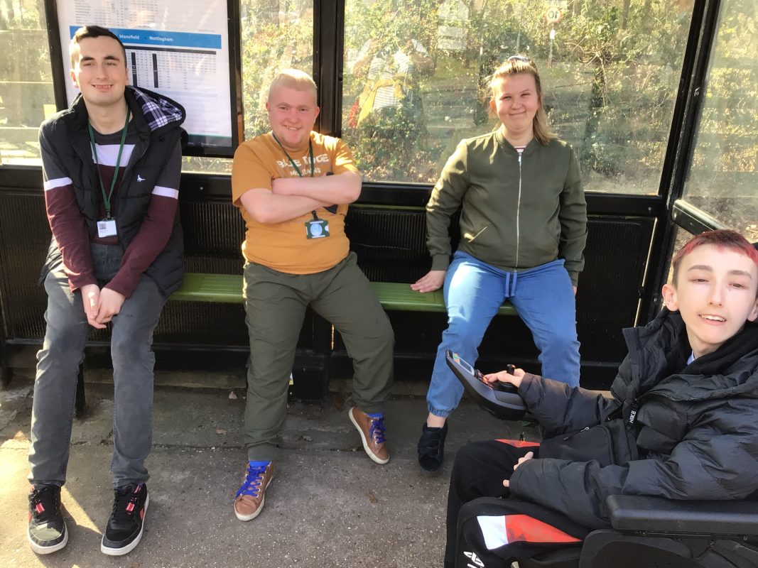 Four people, including one in a wheelchair, are sat at a bus stop waiting for a bus.