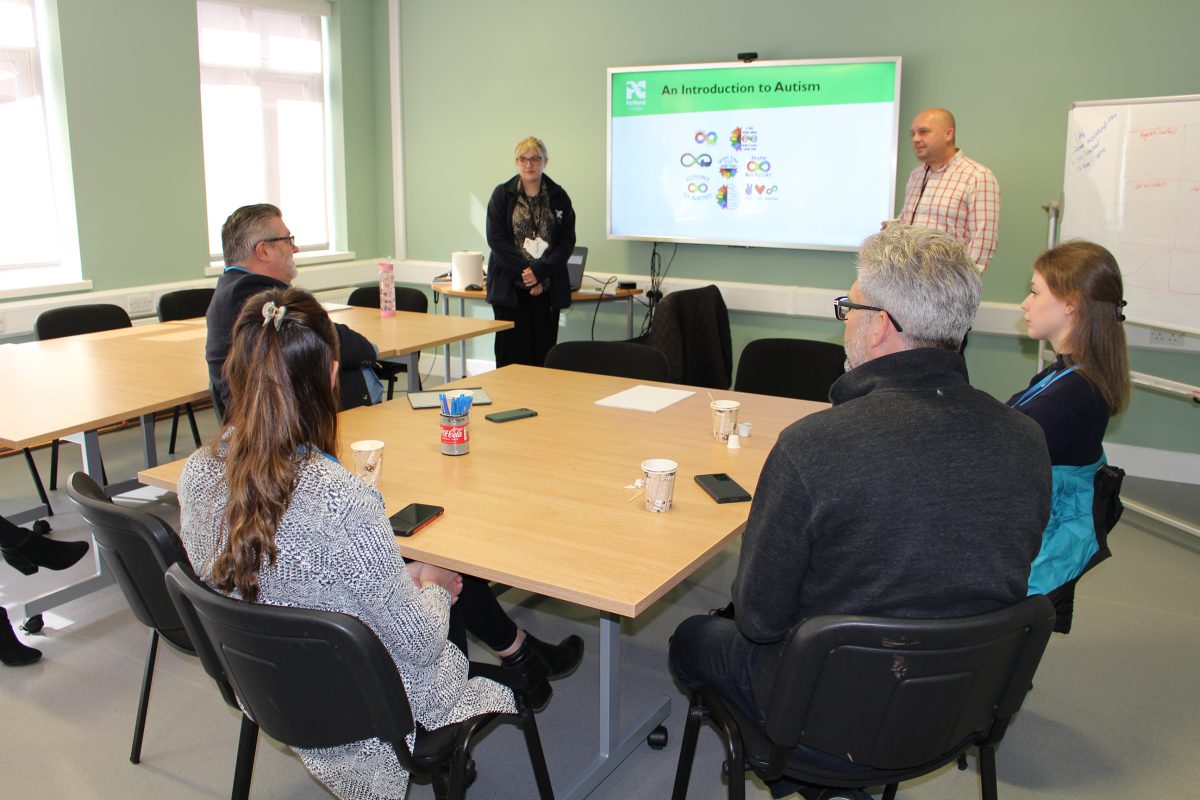 Four people sat around a table watching two more people standing and delivering a presentation.