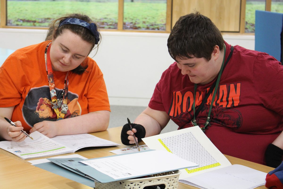 Two people sat working at a desk, girl on right has orange t shirt, boy on right has dark red tshirt