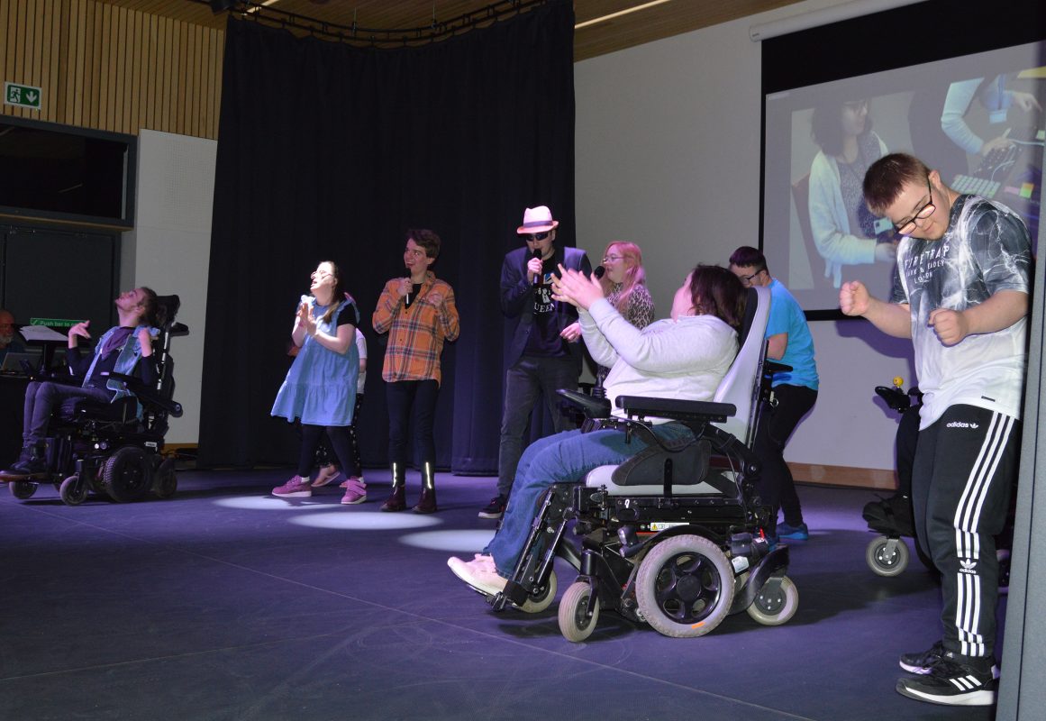 A group of people including two in wheelchairs, singing and dancing.