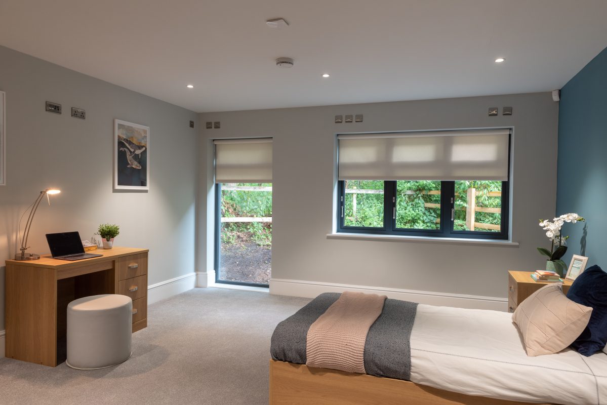 A modern, single bed in the foreground with a floor to ceiling window and other standard window on the wall at the back. A small desk with a laptop and stool at the foot of the bed.