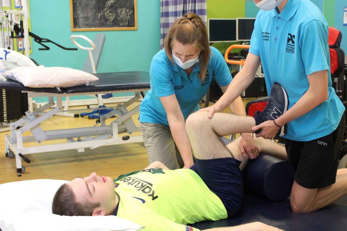 Learner lying down on a physio bed, being helped with leg exercises by two physiotherapists with face masks on