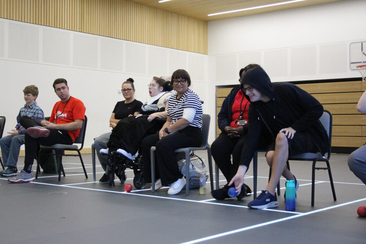 Image of a group of people sat on chairs. One young male is about the release a blue boccia ball