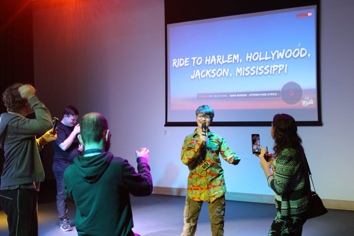 Image of young white male with blond hair singing into a microphone while others dance and take photos