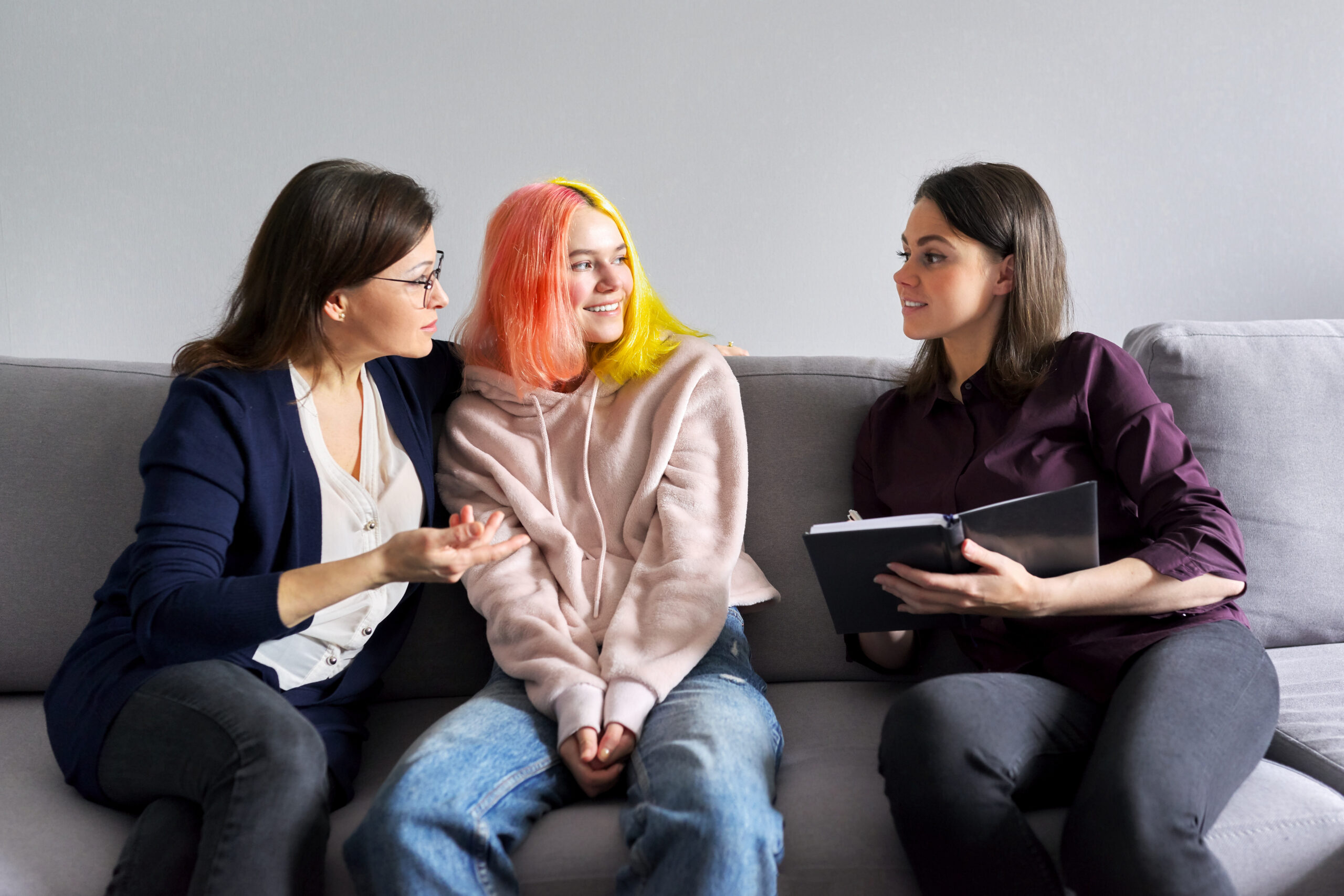 Female teenager with red and yellow hair sat on sofa smiling with mum and assessor