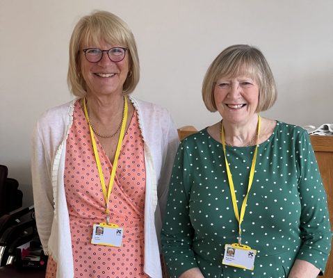 Image of 2 smiling ladies wearing volunteer lanyards with a table of drinks in front of them