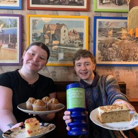 two people smiling and holding out plates of cake and muffins, along with a blue donation tub for Portland College. They’re sitting indoors with colorful framed pictures on the wall behind them, at a cheerful fundraising event.