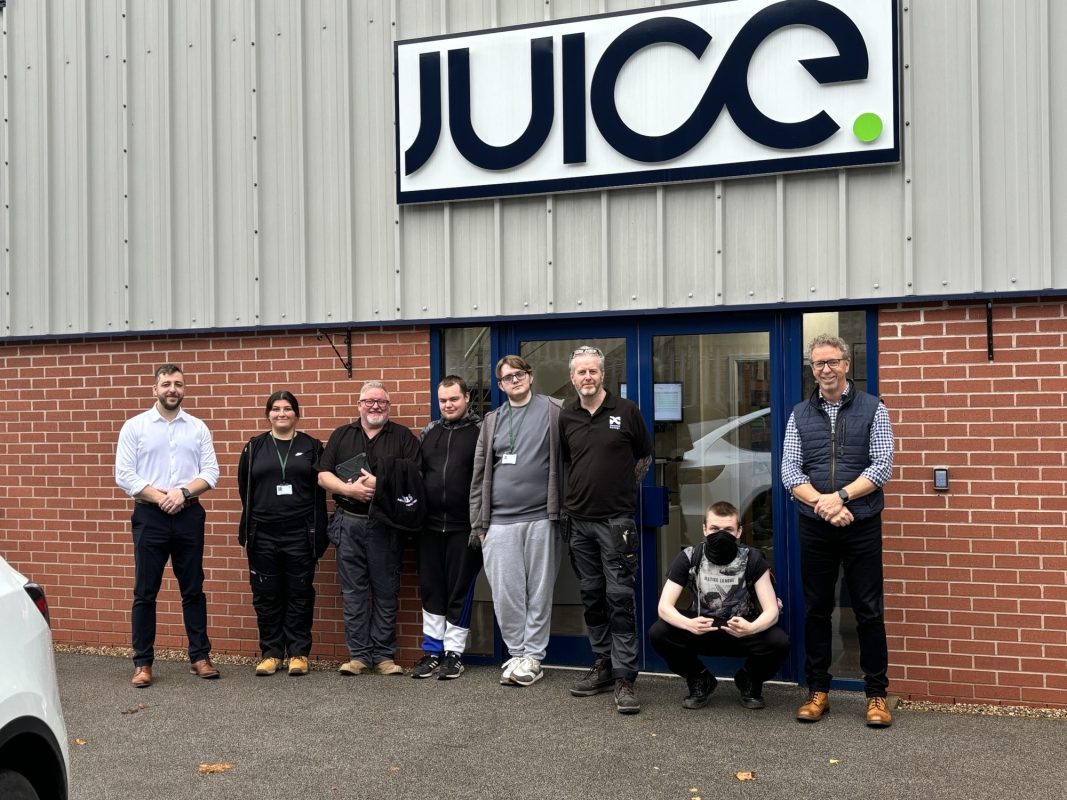 This image shows eight people standing in front of a building with a sign that says “JUICE.” They’re smiling and posing for the photo outside the entrance, which has red brick walls and blue doors.