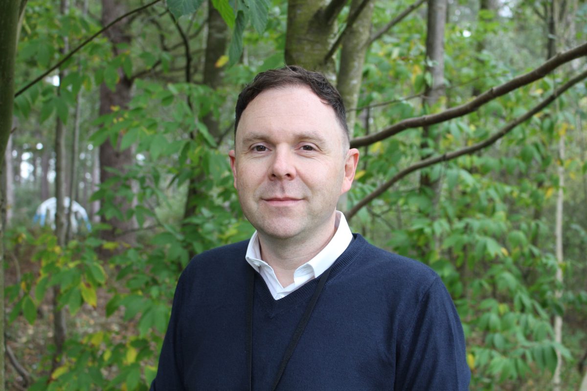 A man with dark hair, dark blue jumper with a white shirt underneath standing in front of some trees looking at the camera.