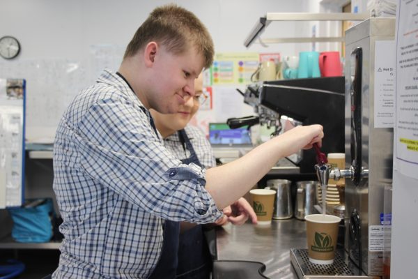 A young man is learning to use a hot water dispenser in a café setting, with guidance from a staff member. Both are wearing aprons and appear focused and engaged. Cups and a coffee machine are visible in the background.