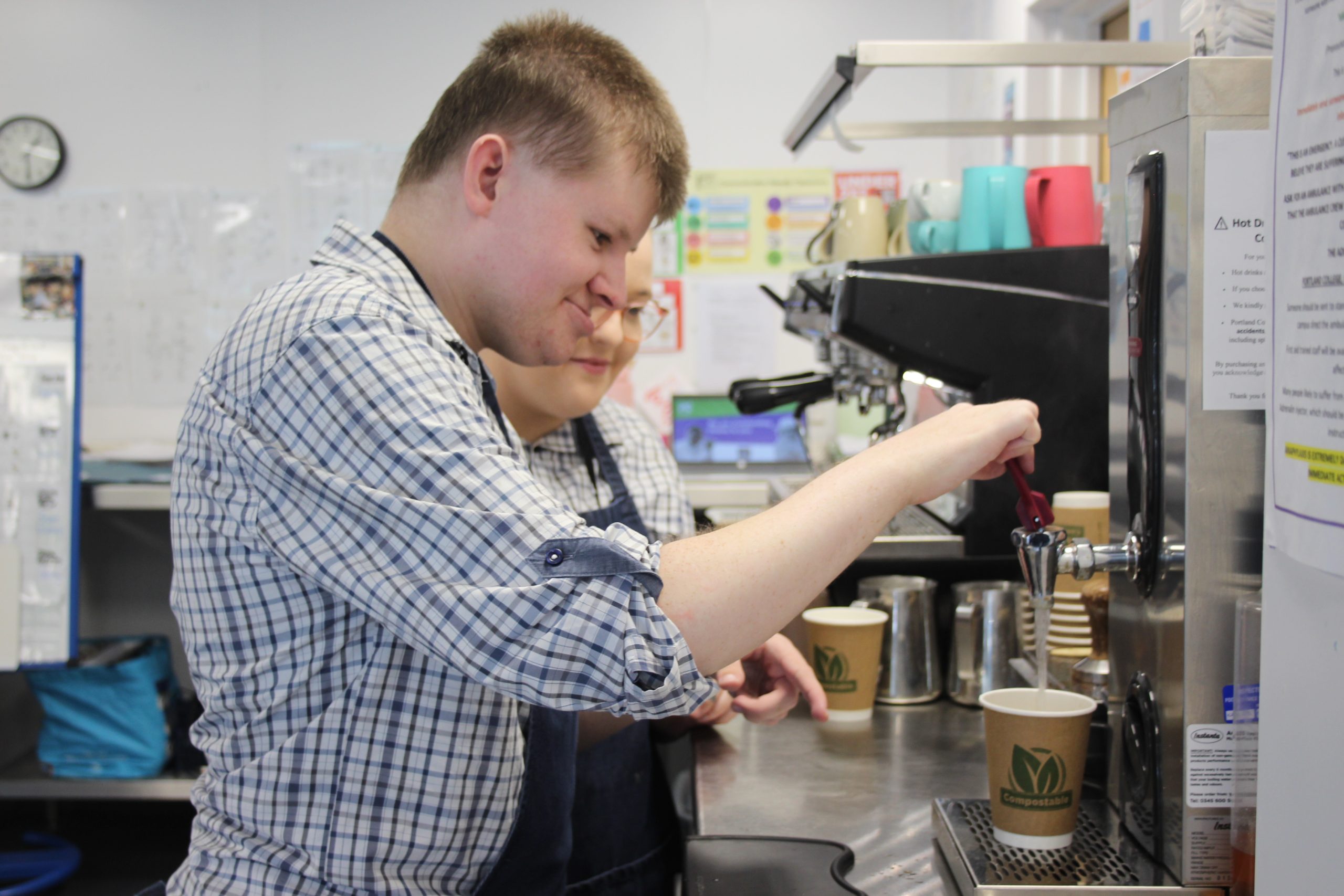 A young man is learning to use a hot water dispenser in a café setting, with guidance from a staff member. Both are wearing aprons and appear focused and engaged. Cups and a coffee machine are visible in the background.