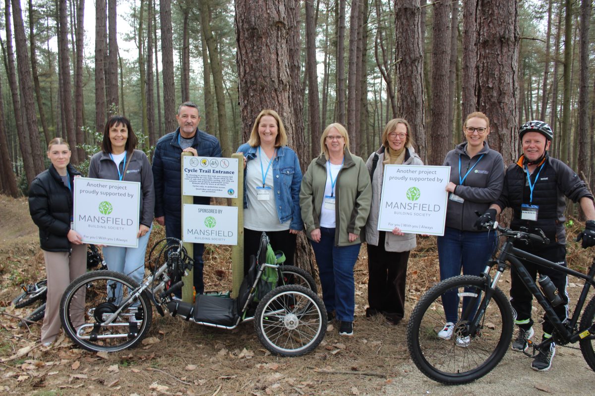 This image shows a group of eight people standing in a wooded area, smiling at the camera. They’re holding signs showing support from Mansfield Building Society, and there are bicycles in front of them, suggesting a cycling project or trail event.