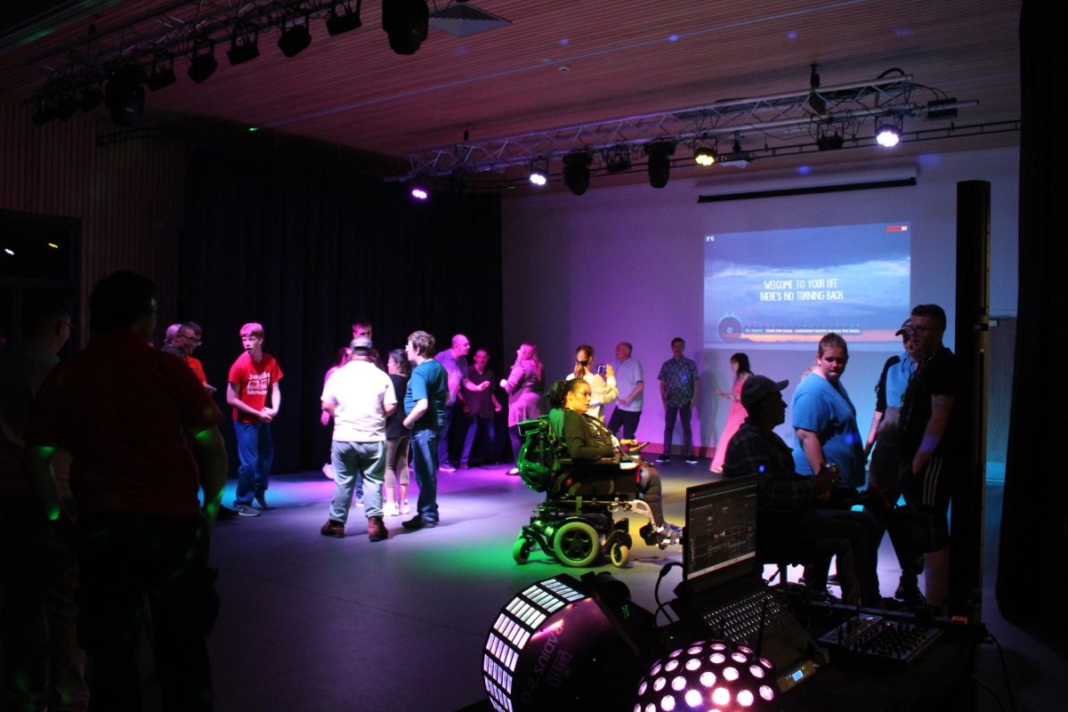 A lively, inclusive dance event with colorful lighting. People of all abilities, including wheelchair users, are socializing and dancing. A screen displays lyrics, and a DJ setup is visible in the foreground.