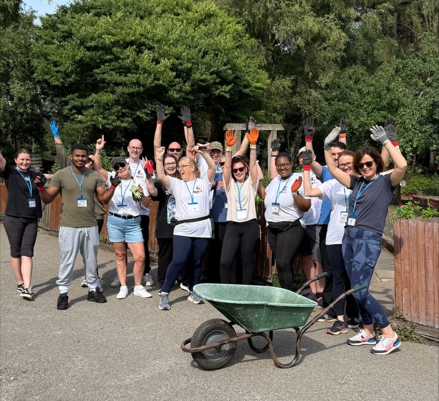 This image shows a cheerful group of people outdoors, raising their hands and smiling for the camera. They’re wearing work gloves and name badges, standing on a path with trees behind them and a wheelbarrow in front