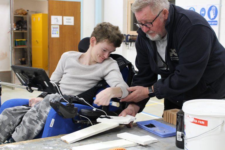 A young man in a wheelchair paints wood with support from an older man. They're working together in a workshop, sharing a positive, hands-on learning moment.