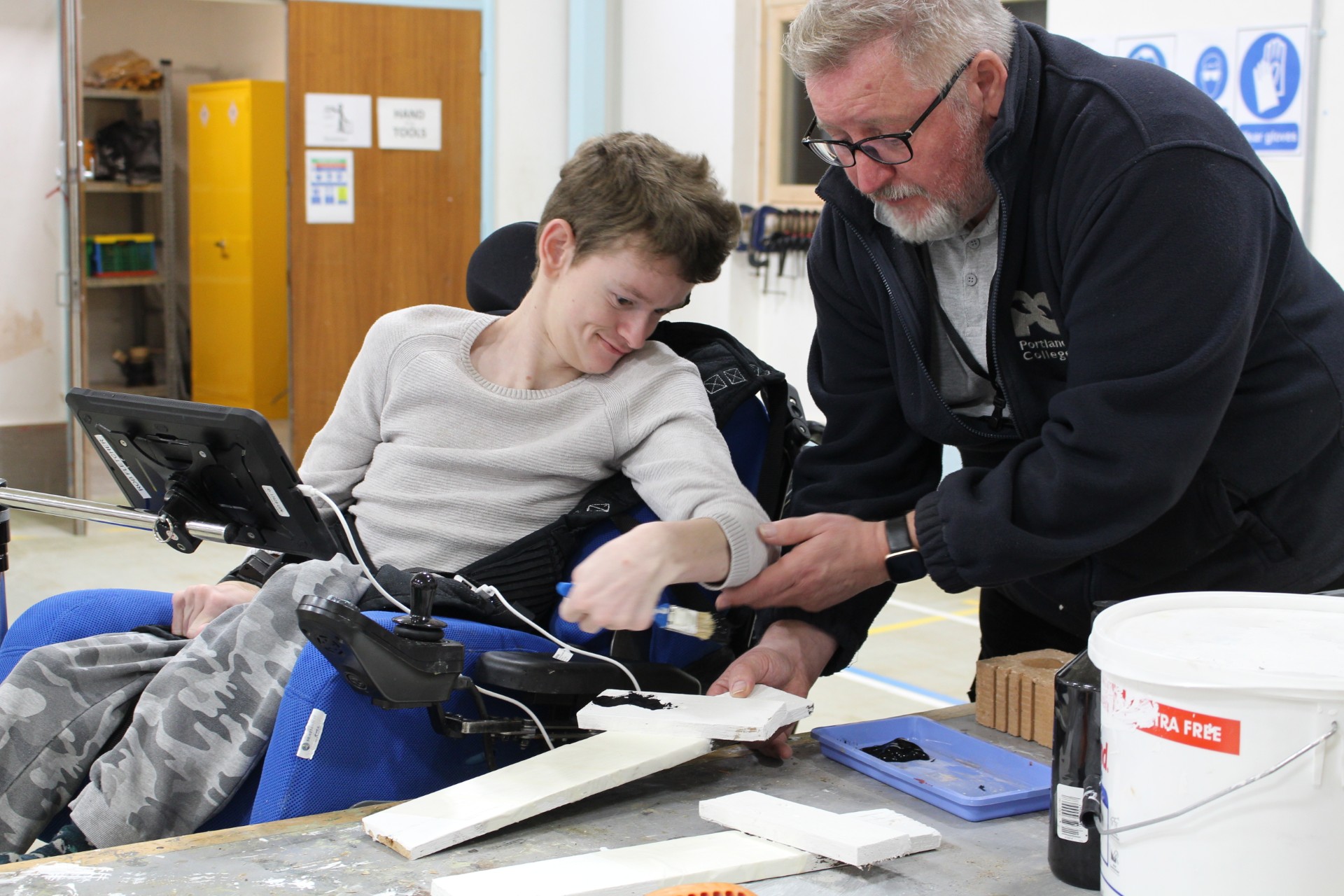 A young man in a wheelchair paints wood with support from an older man. They're working together in a workshop, sharing a positive, hands-on learning moment.