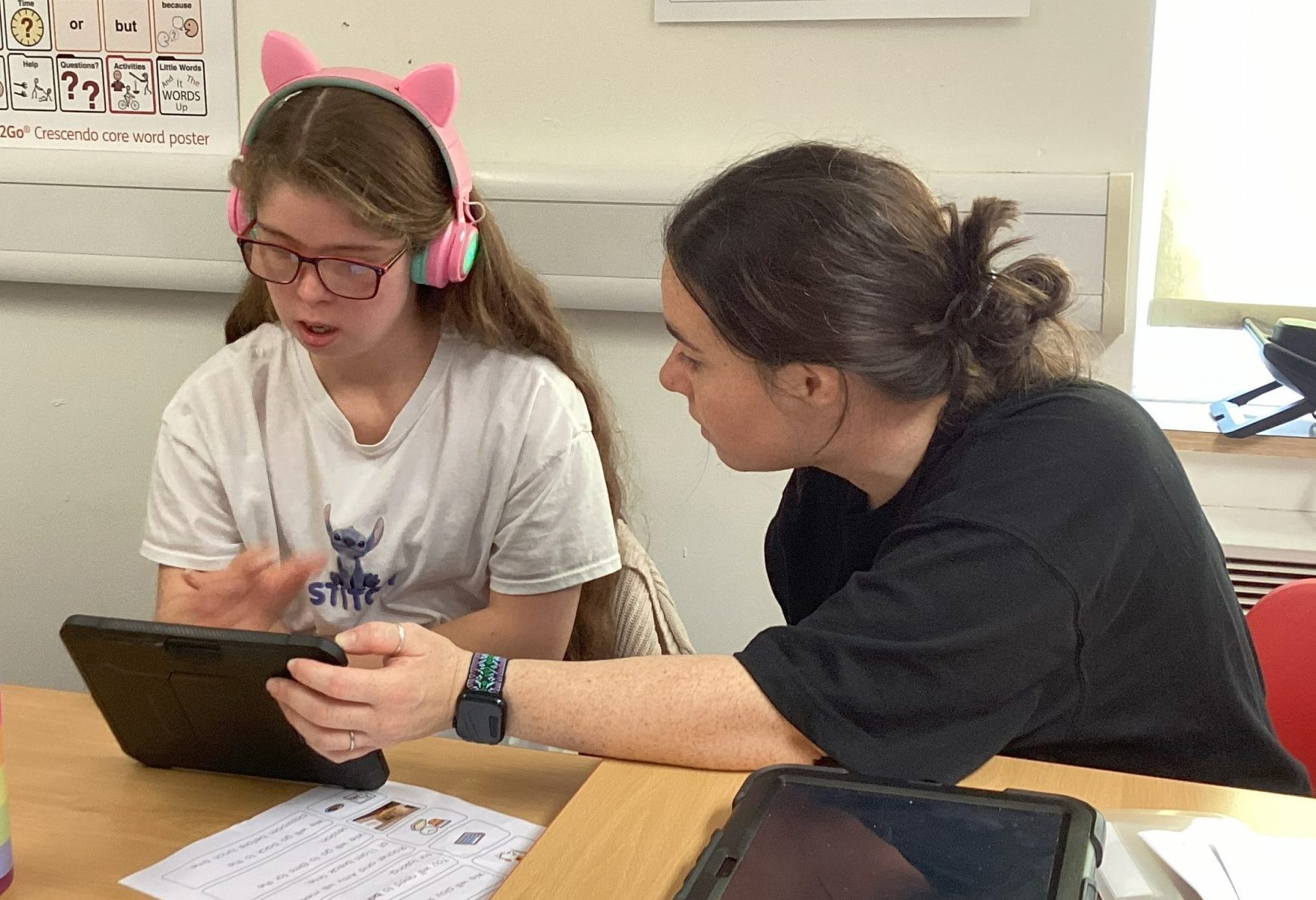 young girl wearing pink headphones and brunette female teacher holding an Ipad