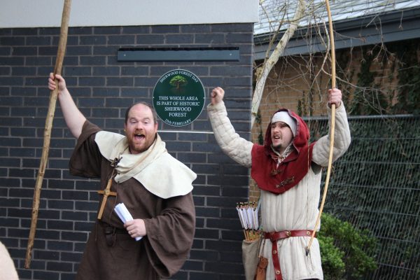 Two people dressed in medieval-style costumes stand in front of a brick wall, holding longbows and raising their arms in celebration. A green plaque behind them reads that the area is part of historic Sherwood Forest. One person holds a scroll, and a quiver of arrows is visible on the other’s belt.
