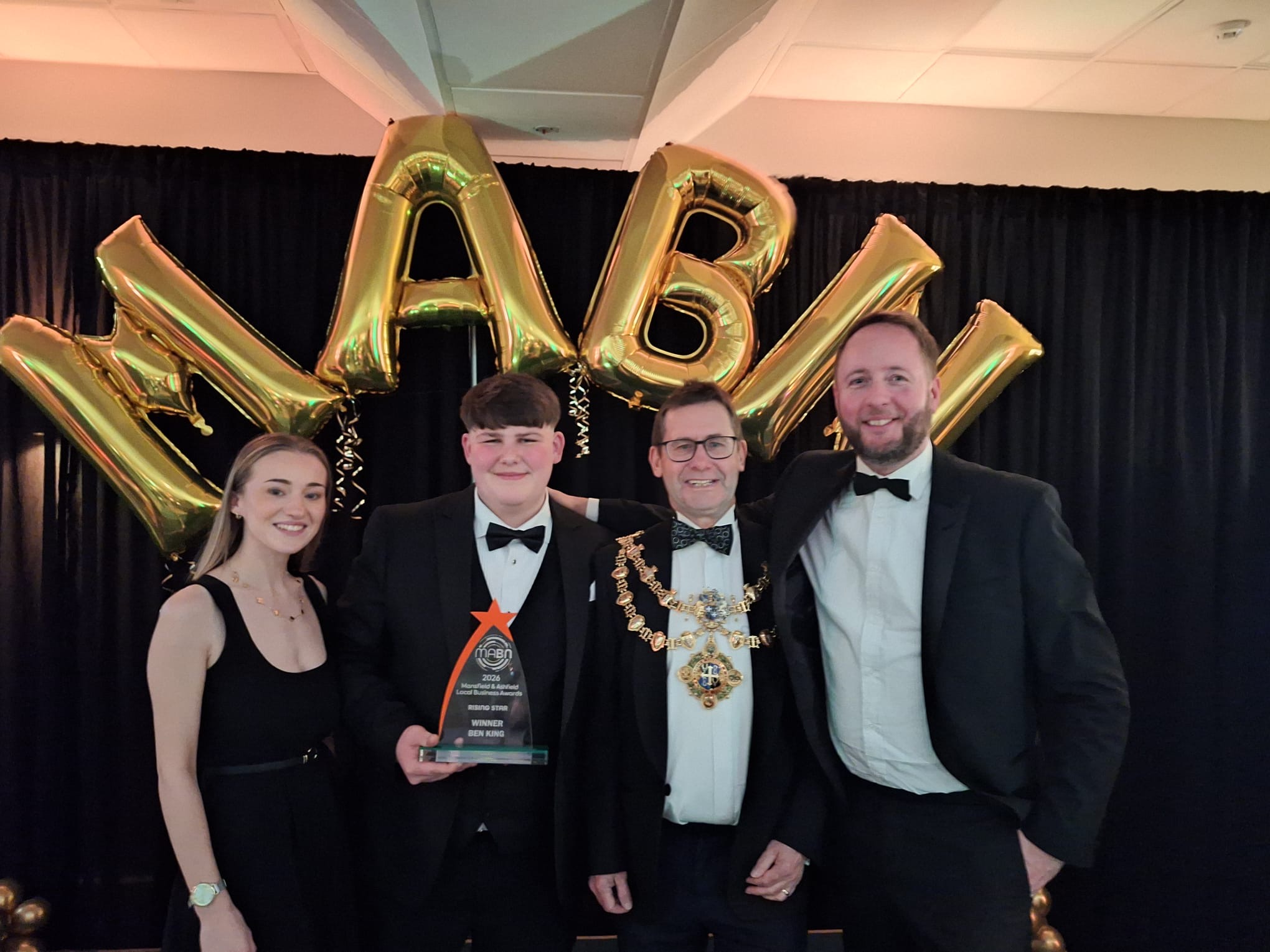 A smiling group photo with three men in black tie suits and one blonde female in a black dress