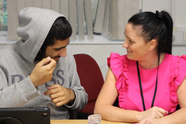 A male in a grey hoodie with the hood up on the left sat at a desk with a woman with a bright pink top, dark hair in a ponytail, looking at each other and smiling.